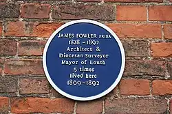 Blue circular Plaque on the side of red brick building with raised white lettering mentioning James Fowler's lifespan of 1828 to 1892 and his residence within from 1869 to 1892