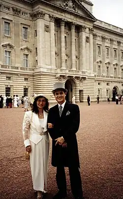A colour photograph of Jason Tsai, alongside his wife Ruth Tsai. The couple are dressed in formal wear: Jason is wearing a black morning suit with a grey top hat and matching grey necktie and pocket handkerchief; his wife Ruth is wearing a full-length white dress, matching white jacket and white hat. The couple are standing in the red-gravelled courtyard of Buckingham Palace – the central London official residence of Queen Elizabeth II, the reigning monarch of the United Kingdom of Great Britain and Northern Ireland. In the background are other unidentifiable people in similar attire.