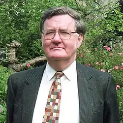 Griffin, an older man with glasses, sideburns and a stern expression, wearing a formal suit. He is photographed on a summer's day in a college garden.