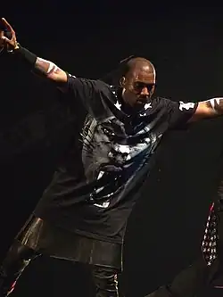 A man holding a microphone in his right hand whilst on stage. He wears a black T-shirt with a tiger-like face on it, tight leather pants, and a kilk.