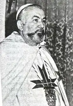 Black and white photo of Origas dressed in Templar garb, wearing white robes with a Templar cross and medallion