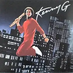 A man in a red jumpsuit is shown in mid-air holding a saxophone in his right hand, over a backdrop of a city at night with lit-up buildings. At the top right is written "Kenny G".