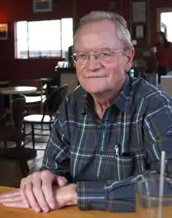 Color photographic portrait of an elderly man with greying hair and mustache, wearing glasses, sitting with hands crossed at a table with a pen tucked in his pocket