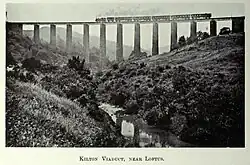 A black and white image of a steam train crossing a tall viaduct