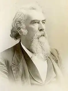 A man with white hair and a white beard and mustache, facing right. He is wearing a white shirt, black vest, and black jacket