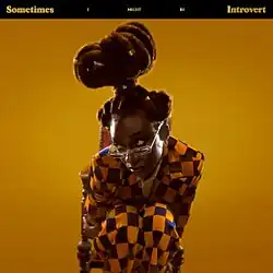 Simz hunches forward in her wooden chair and looks shyly over her glasses. Her hair is pulled up in a bun high above her head and she wears a brightly checkered suit.