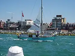blue-hulled two-masted yacht with mainsail partly set passing a rocky shoreline with buildings beyond