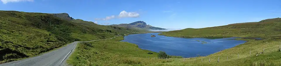 A blue body of water sits beneath a blue sky surrounded by green moorland. A road to the left travels along the lakeside leading towards a small patch of mist and some low hills in the distance.