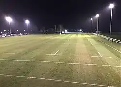 An image overlooking the main pitch of Long Buckby RFC with floodlights on at night.