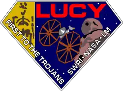 A diamond-shaped crest houses artworks of the Lucy fossil at left, the Lucy spacecraft at center, and an artist's impression of a Jupiter trojan. The word "Lucy" is written in a large, bold red font at the top right corner, while the words "First to the Trojans" and "SWRI · NASA · LM" are written in a smaller white font across the bottom edges of the diamond-shaped crest.