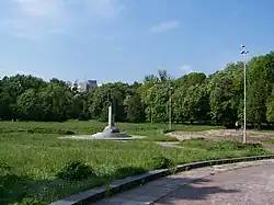 A green park with a statue in the middle of the picture. The statue has three small steps up to its base, which is a half football. There is then a column leading up to a bronze-brown falcon statue. It is difficult to see the details but the falcon is on top of a football as well.
