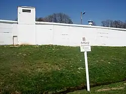 At the top of a grassy slope, there is a white concrete wall; treetops, a lamppost and the top of a guard tower are visible over the top of the wall. In the foreground, there is a white sign saying "Achtung! Bachmitte Grenze".