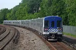 An eight-car passenger train, silver on the sides and blue on the front, on a curved stretch of track with a third rail