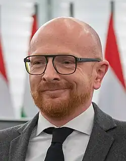 close-up of Mads Brügger wearing a grey suit jacket, white shirt, and black tie, looking right of camera with a one-sided smile