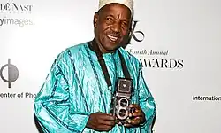 The image features Malick Sidibé, a celebrated Malian photographer, at the 24th Annual ICP Infinity Awards. He is dressed in a blue traditional Malian attire known as Bazin. Holding a camera in his hands, he smiles warmly, embodying a spirit of creativity and passion for photography. The setting reflects the significance of the event, celebrating artistic achievements in the field of photography.