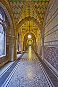 Gothic first floor corridor, Manchester Town Hall, (the corridors higher in the building are much plainer) showing use of ceramic wall tiles of different colours making a geometrical pattern, the upper walls are plain buff terracotta with horizontal lines of blue tiles running through (this was before faience became available), the floor is terrazzo with patterned mosaic borders, the vault painted with geometrical multi-coloured designs, also note the two decorative cast-iron grilles running along the floor, these cover the heating pipes