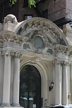 The entrance on 108th Street, which is through a set of glass and iron double doors. The top of the archway contains a keystone. On either side of the arch are two protruding Ionic columns, which stand in front of two Doric pilasters. Atop the entrance pavilion is a cornice with brackets, which rises above a segmental arch.