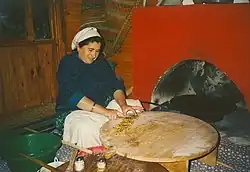 Traditional tableside preparation of gözleme in a restaurant near Antalya. A gas-powered sac griddle can be seen inside the red oven to the right, with two thin wooden rolling pins ("oklava") and pastry brushes used to prepare the dough to the lower left. The cook is seen cutting up the finished gözleme on her pastry board table to prepare for final serving.