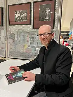 Mark Edwards wearing a black coat, sitting at a desk during a book-signing, looking at camera from profile with slight smile