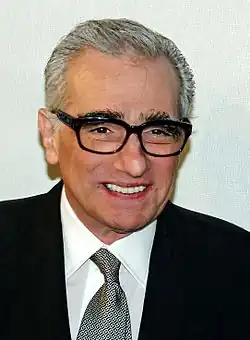 A Caucasian man in his 70s smiling. He is wearing black framed glasses and a tie with a black suit over a white shirt. Behind him is a white background