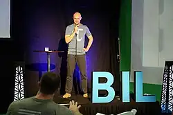 Michael Hartl standing onstage holding a microphone with the letters BIL in the foreground answering questions after his Tau Talk at the BIL Conference