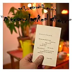 A man's hand holding the Joanne Kyger poem "Night Palace" in the foreground. Background is of a house illuminated by an orange light.