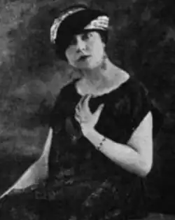 A white woman, seated, wearing a turban-style hat and a loose-fitting black dress with bare arms; she has one hand on her chest