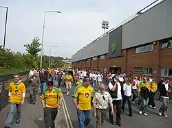 A group of people wearing predominantly yellow football shirts, walking along a road beside an association football stadium