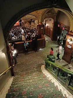 View down the northern stairs from the first balcony to the orchestra level, behind the right side of the auditorium. The landing at the bottom of the stairs has a vaulted ceiling.
