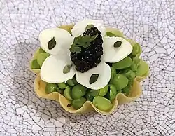 A clamshell-shaped pastry base filled with green beans. A white flower made of white chocolate sits on top; covered with a spoonful of black beads of caviar. It is garnished with small leaves.