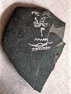 Piece of dark-grey stone bowl inscribed with white hieroglyphs, one of which shows a boat on a river