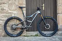 A grey 2020 Orbea full suspension mountain bike with a telescoping suspension fork and a four-bar linkage rear suspension in front of a buildings wall and door.