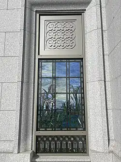Stained glass depicting various water elements and bullrushes, against a blue sky background. A metal frame outlines the glass and above the stained glass depicts a pattern of nine cherry blossoms. Masonry from the temple walls hold the window in place.