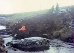 Two British soldiers stand in a shallow river, examining an overturned vehicle. Two other soldiers watch from the riverbank.