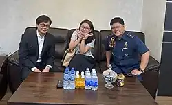 Guo sits on a couch with two men on either side of her. Guo is smiling at the camera while making two V signs.