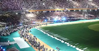 A portion of the stadium with stands full of people, a large artificial tree is on the right side of the image. A group of people are walking together on the stadium floor