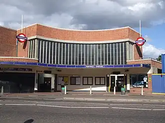 A red-bricked building with a rectangular, dark blue sign reading "PERIVALE STATION" in white letters all under a light blue sky