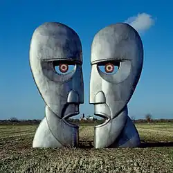 A colour photograph of two large silver-grey iron sculptures of opposing silhouetted faces. The sculptures are standing in a brown wheat field with a blue sky behind them.