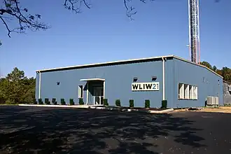 A one-story blue metal building with a W L I W logo next to the base of a tall broadcasting tower. In comparison to the prior photo, the building has been reshaped in the front and is larger, with new landscaping.