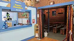On the left, a sign reads, "Welcome to Puppet Showplace BOX OFFICE." Below is a light blue wall with an opening and counter. Puppets are visible on the counter, through the opening, and near the sign. On the right, an opening in a brick wall leads to the theater. Colorful curtains frame the opening. In the theater, there are benches with light blue cushions.