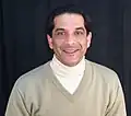 A man viewed from the chest up, wearing two sweaters at once. The outer layer is a beige v-neck knit sweater with long sleeves and a rib knit neckline. The inner layer is a white turtleneck sweater.