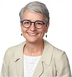 Portrait of a woman in head-and-shoulders, directly facing front. She is smiling, wearing spectacles and a light, neutral-coloured jacket over a white top. She has short, grey hair.