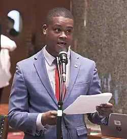 A black man in a blue suit with red tie and white shirt standing behind a microphone holding two sheets of paper