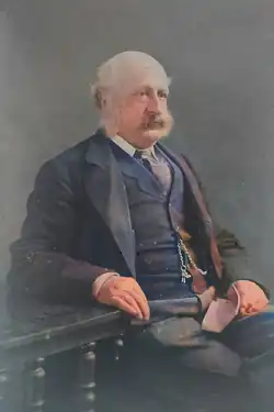 A photograph of an old man with white hair and a white beard. He is sitting on a chair facing sideways to the viewer, and his gaze is not directly towards the viewer either. He is wearing a navy three-piece suit.