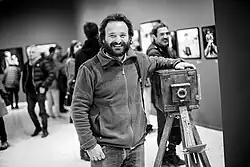 Black and white photograph of Abd, leaning against an old camera, in a museum