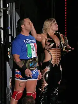 A man and a woman pose together. The dark-haired man is standing on the left and is wearing a blue T-shirt with the Italian flag and the word 'Santino' written upon it, blue wrestling tights, and red kneepads. He is wearing a gold wrestling championship around his waist. The blond-haired woman is standing on the right, wearing a black catsuit with the sides of the torso removed to bare part of her midriff. She is holding a wrestling championship upon her left shoulder and has her right arm outstretched.