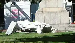 A white marble fallen statue depicting Robert Falcon Scott, on the ground in a small urban park