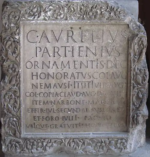 Epitaph displaying apices and long i. Nimes, 1st–2nd century CE. C(aivs) · Avrelivs Parthenivs órnámentꟾs · dec(vrionalibus) honórátvs · col(oniae) · aug(ustae) nemavsꟾ ꟾiiiiꟾvir · avg(vstalis) col(onia) · cópia · clavd(ia) · avg(usta) · lvgvd(vnensis) item · nárbóne mártio et · fir(ma) · Iv́l(ia) · secvnd(anorum) · aravsióne et · foro · ivliꟾ pácáto vbꟾqve · grátvitꟾs · honóribvs