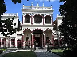 Through gardens and a fence, a two storey building can be seen. The front door is protected by Chinese guardian lions. The facade has Victorian columns and features.