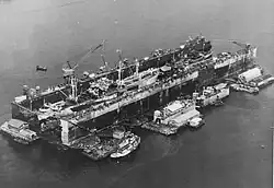 A floating drydock containing ships. It is surrounded by floating barges with workshops and a tugboat.